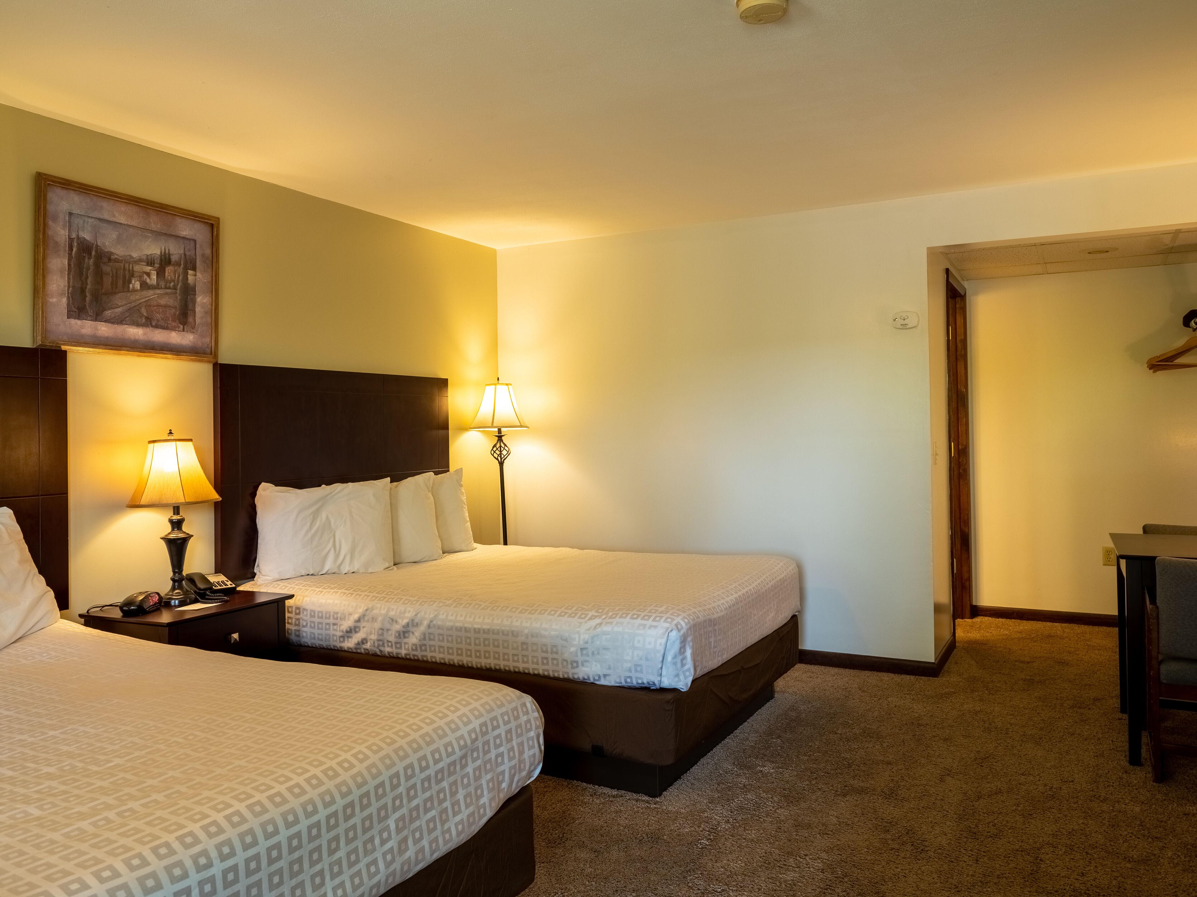 A hotel room with two beds, a nightstand with a lamp, a painting on the wall, and a door leading to another room.