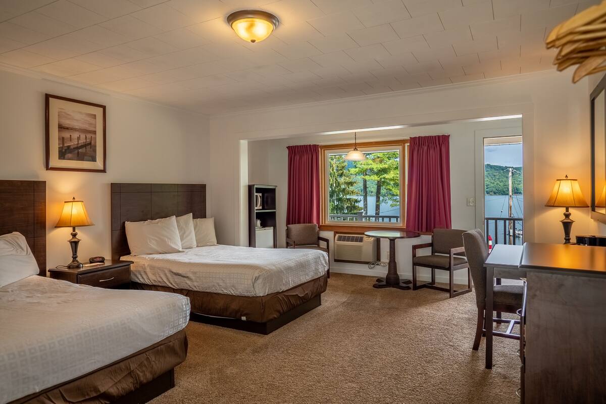 Ein Hotelzimmer mit zwei Betten, einem Schreibtisch, einem Stuhl, einem Fenster mit Blick auf Bäume und Wasser sowie einer Deckenleuchte.