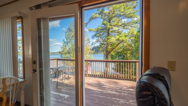 A room with a sliding glass door leading to a wooden deck with a view of a lake and trees. - Lake 'N Pines Motel (Cooperstown)