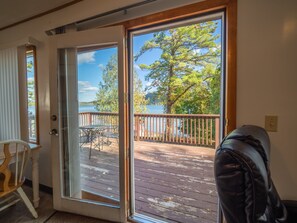 A room with a sliding glass door leading to a wooden deck with a view of a lake and trees. - Lake 'N Pines Motel (Cooperstown)