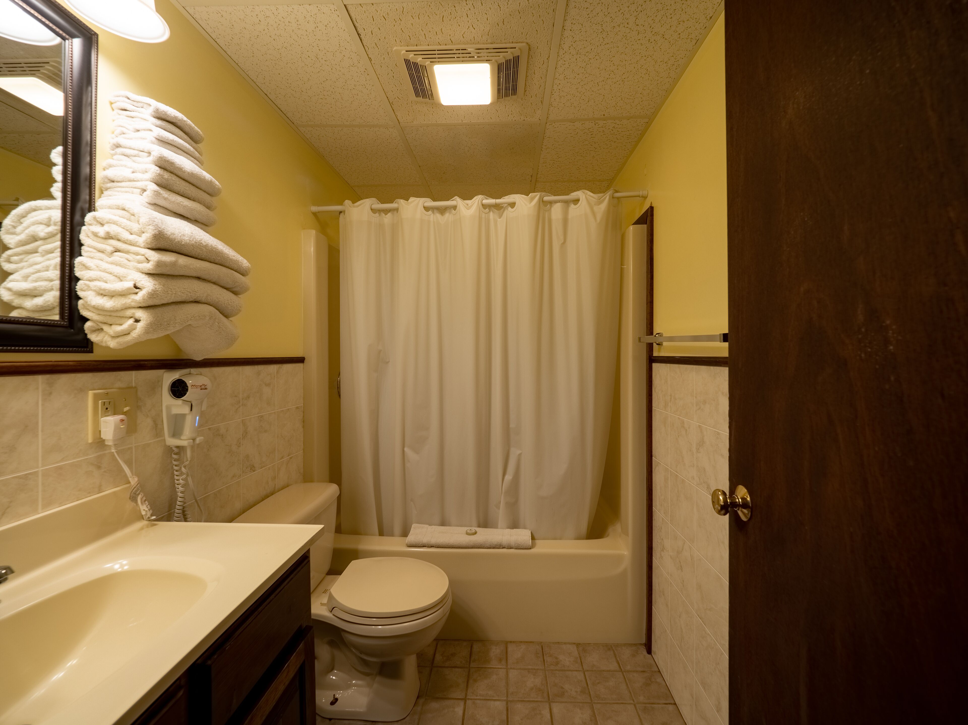 Een kleine badkamer met toilet, wastafel en bad. Handdoekrek met opgevouwen handdoeken, een spiegel en een douchegordijn.