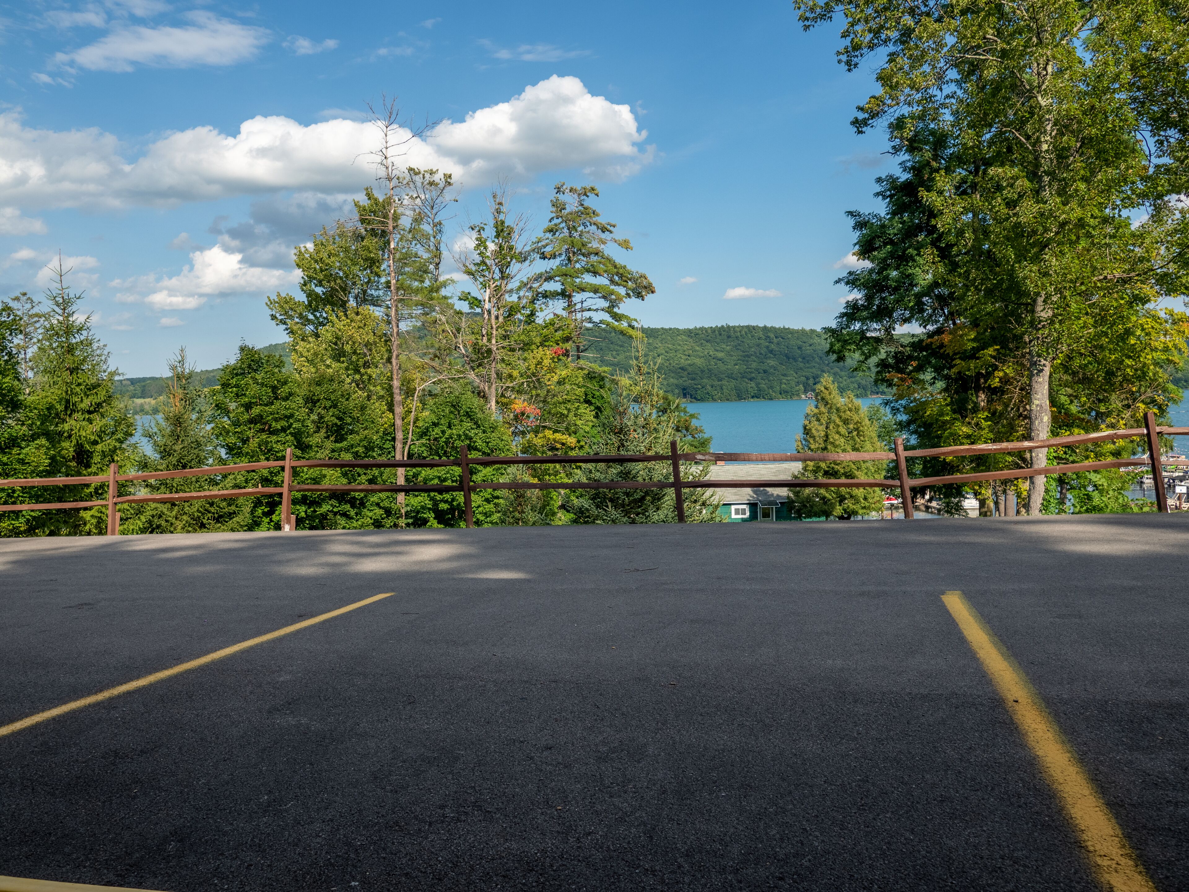 Un'area di parcheggio asfaltata con recinzione in legno, circondata da alberi e con vista su un lago e montagne.