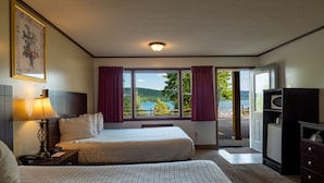 A hotel room with two beds, a desk with a lamp, a television, and a view of a lake and mountains. - Lake 'N Pines Motel (Cooperstown)