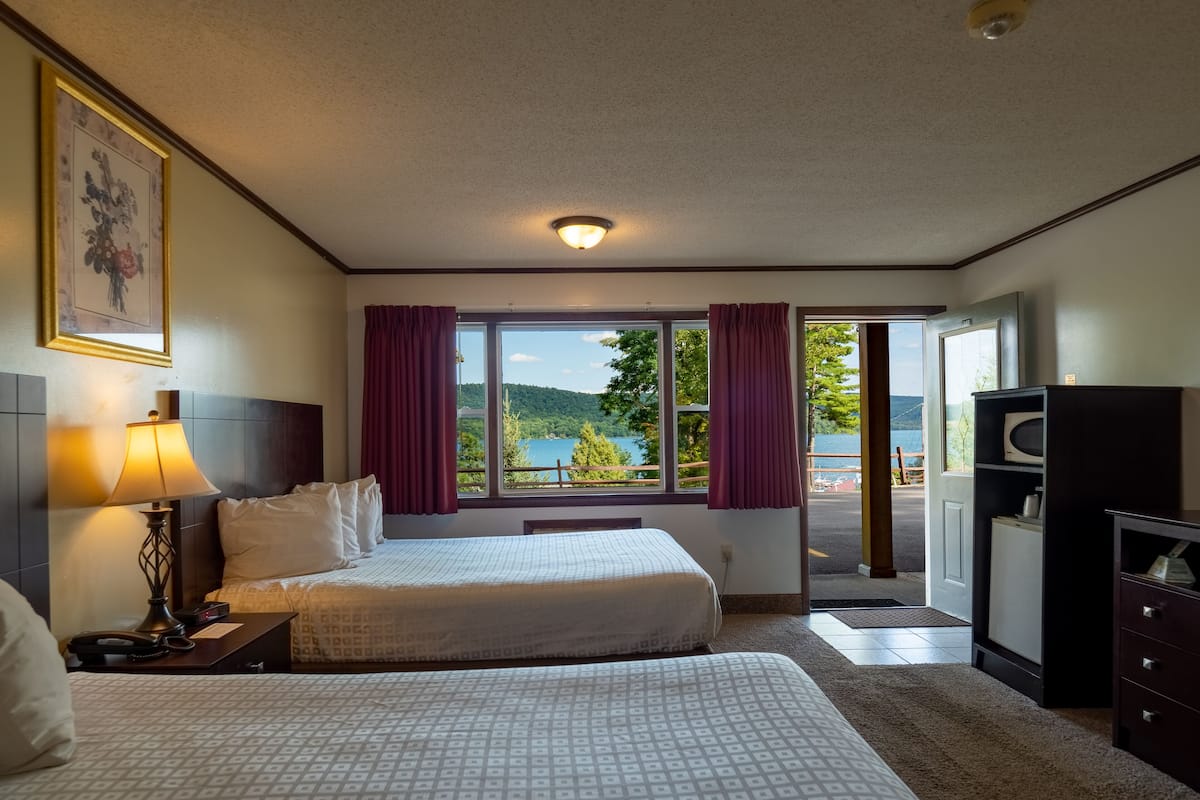 Ein Hotelzimmer mit zwei Betten, einem Schreibtisch mit Lampe, einem Fernseher und Blick auf einen See und Berge.