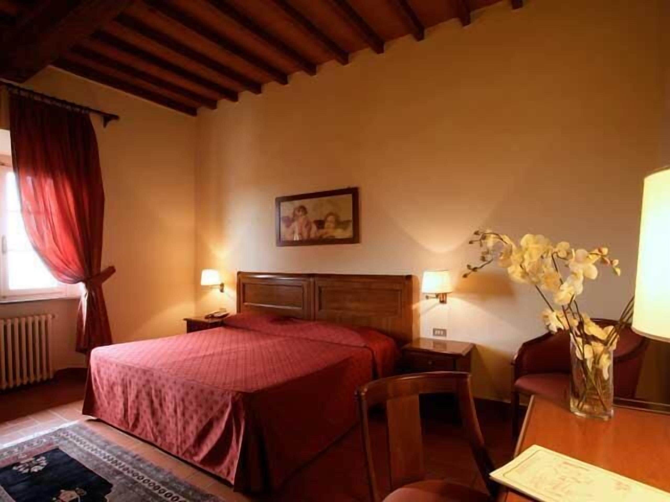 A hotel room with a bed, two bedside tables, a chair, a table, a vase with flowers, a painting on the wall, and a window with curtains.