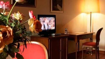 Habitación de hotel con televisor sobre un soporte de madera, un escritorio con silla y un florero con flores.