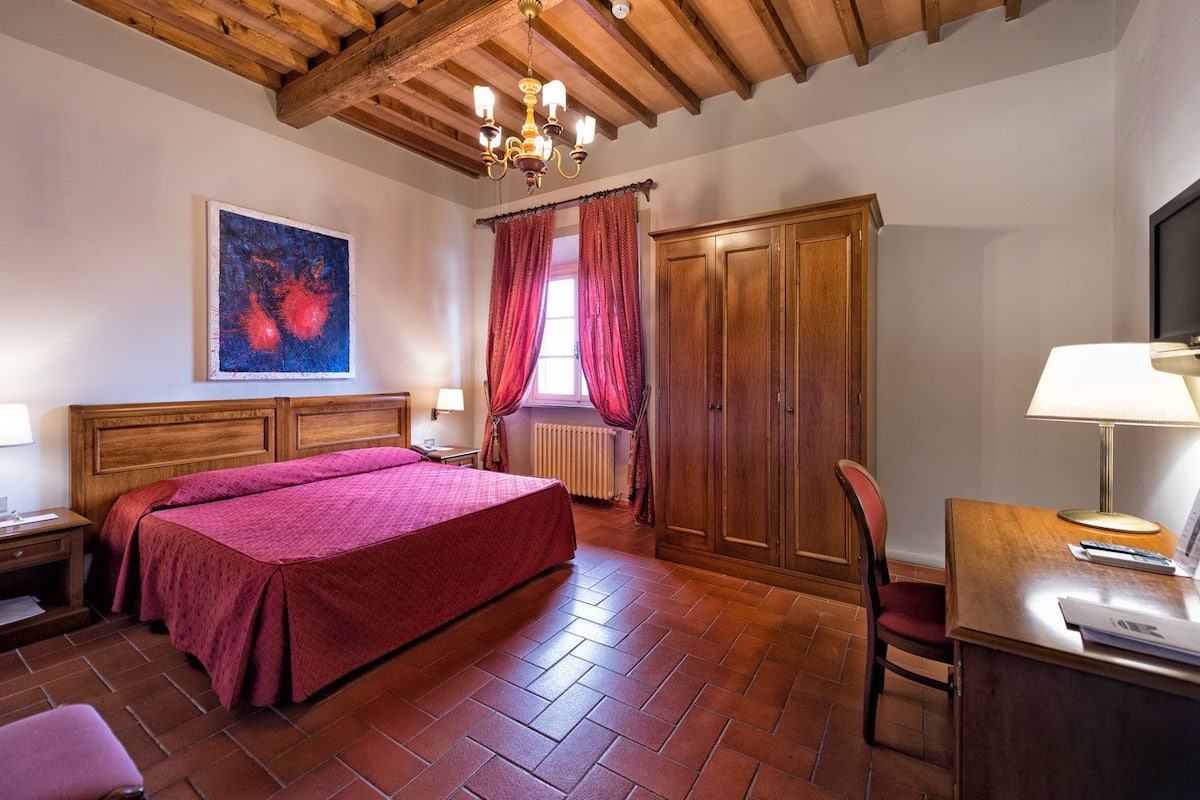 A hotel room with a large bed, wooden wardrobe, and a desk with a lamp.