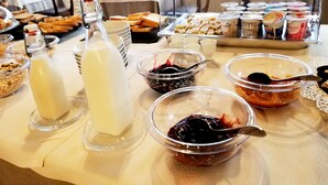 Mesa de buffet com diversos alimentos, incluindo pão, geleias e um dispensador de leite.