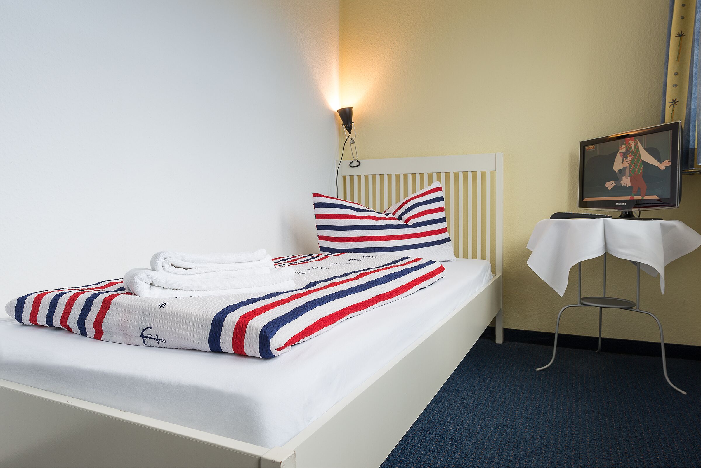A neatly made bed with striped bedding, a bedside lamp, a small table with a white cloth, and a television on a stand.
