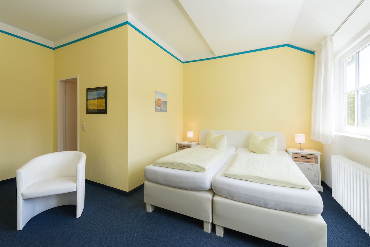 Ein Hotelzimmer mit Doppelbett, einem Sessel, einem Nachttisch mit Lampe und einem Vorhangfenster.