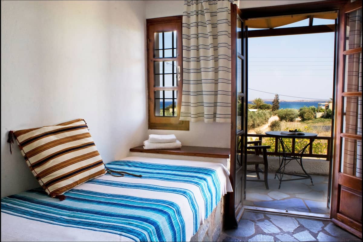 A bedroom with a bed, a window with curtains, a small table and chair outside, and a view of the landscape.