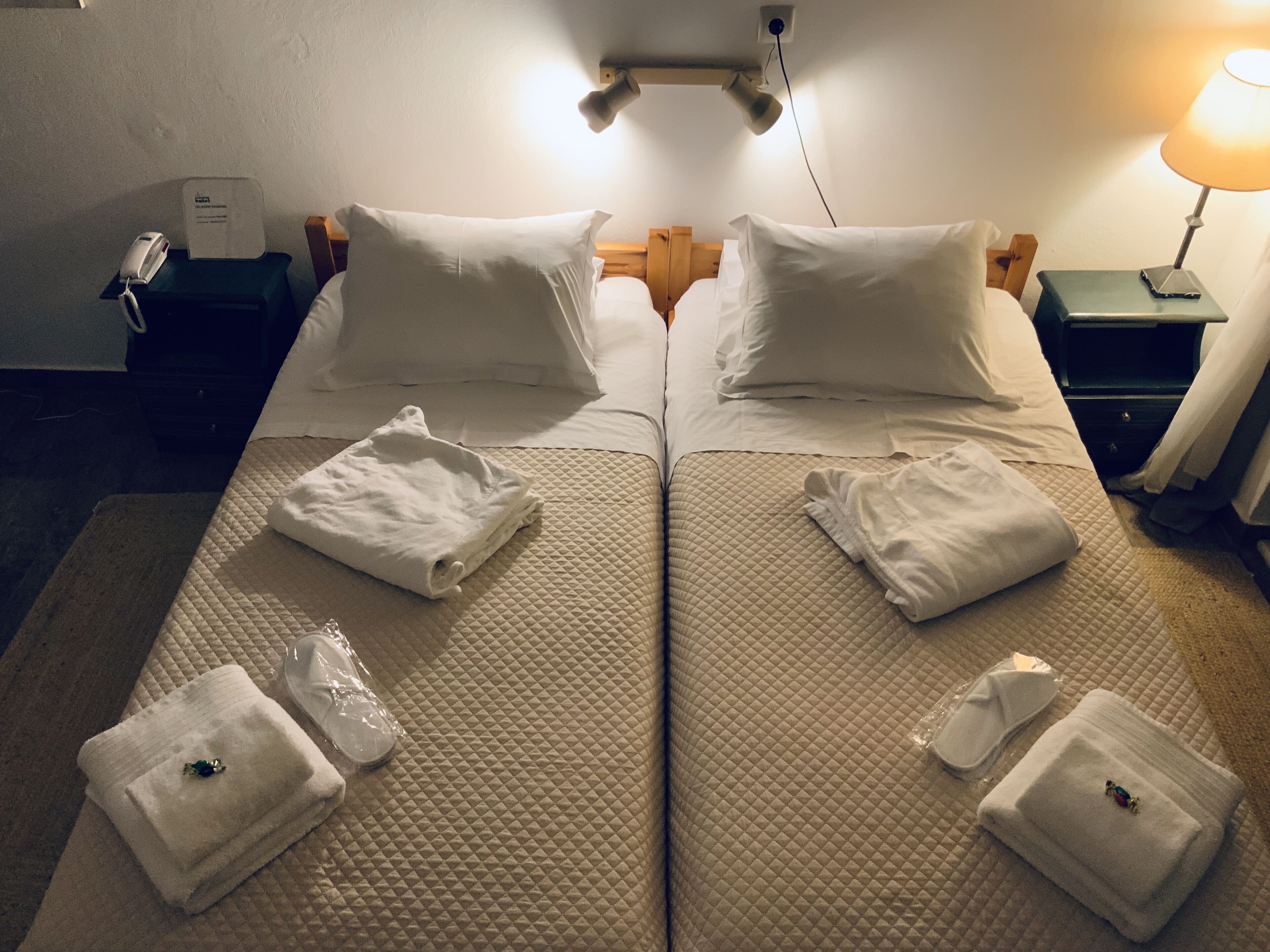 Habitación de hotel con dos camas, cada una con ropa blanca y almohadas. Hay toallas y una mesita con una lámpara.