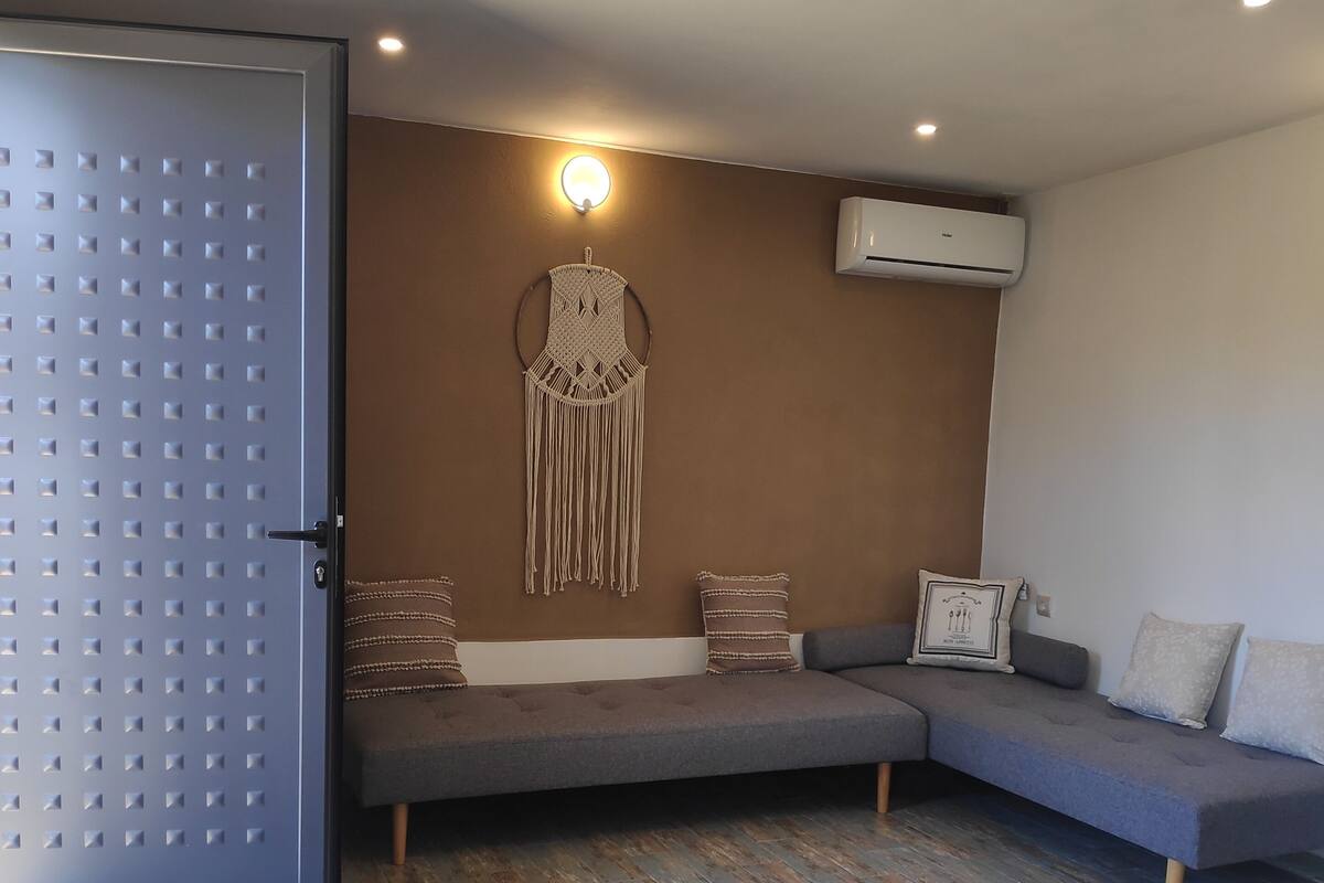 Una sala moderna con un sofá seccional gris, un colgador decorativo de macramé y un equipo de aire acondicionado.
