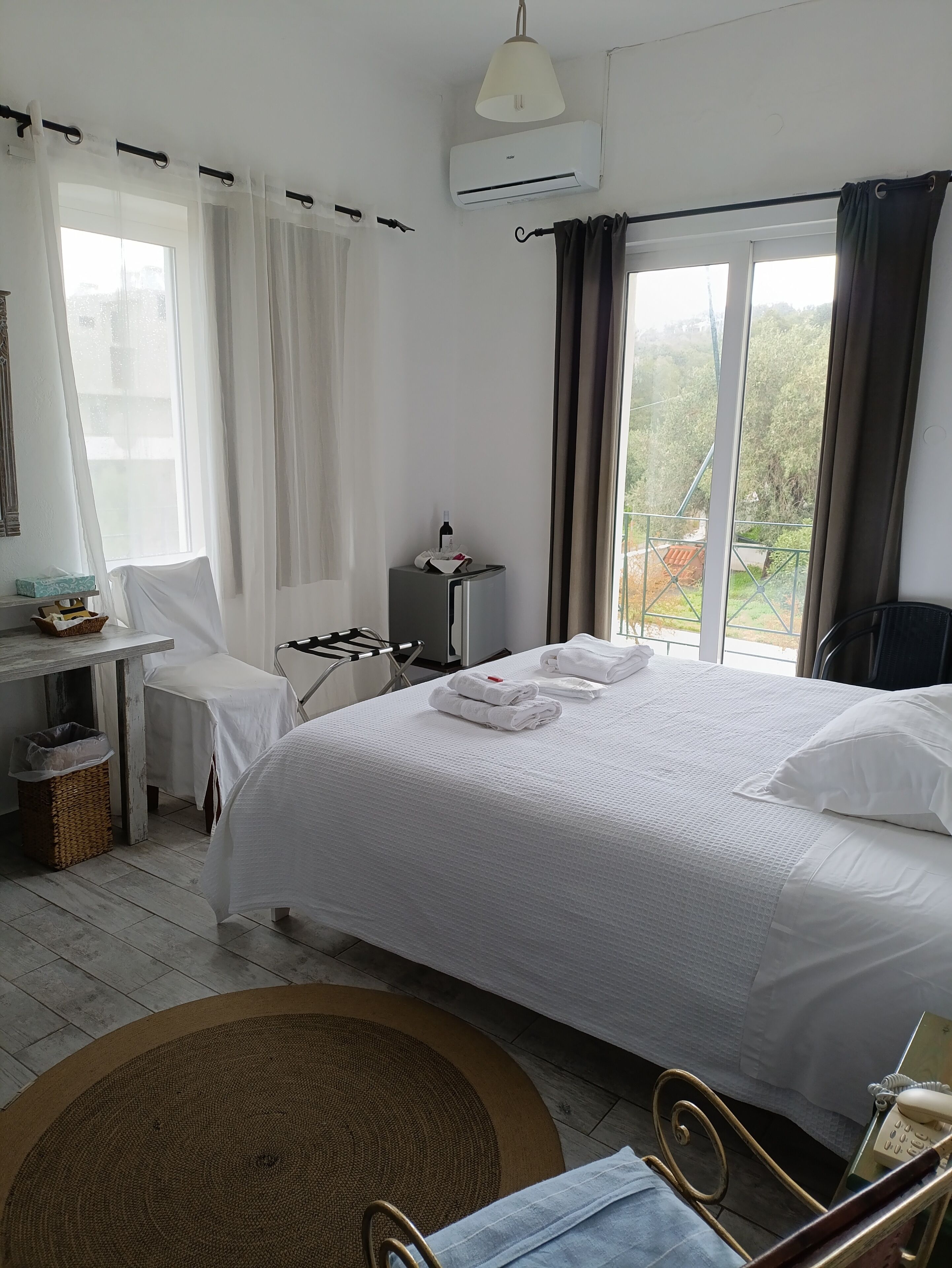 A hotel room with a bed, a desk, a chair, a small table, a trash bin, and a view of the outdoors.