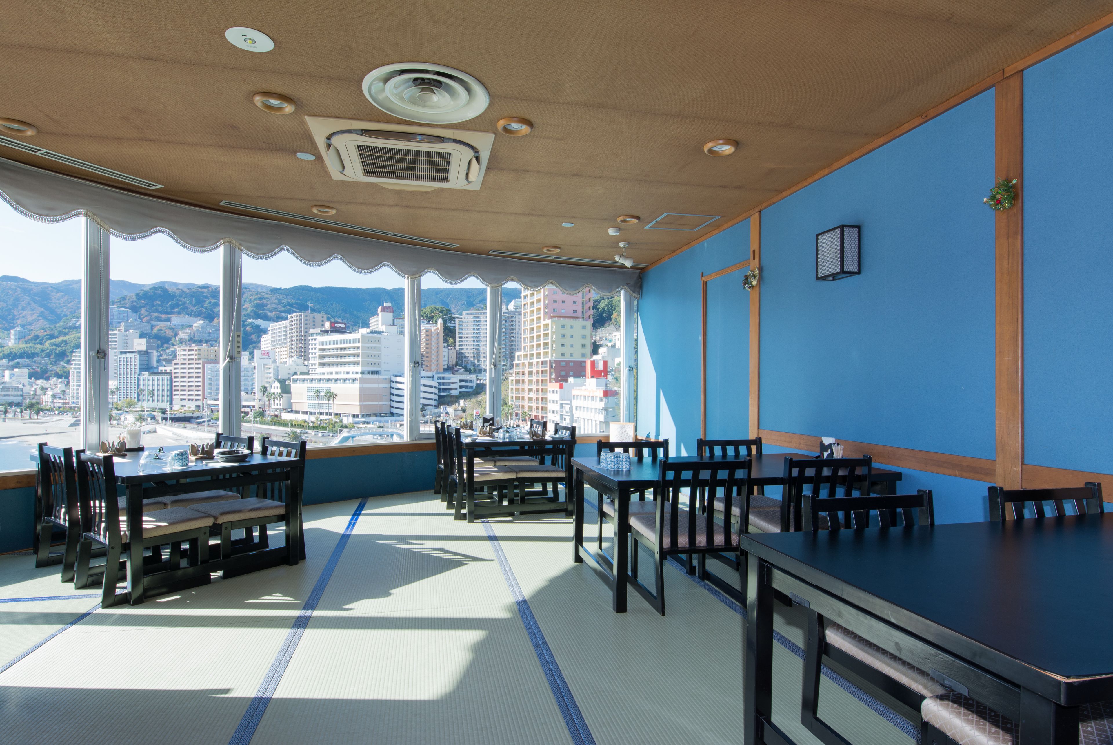 A restaurant with tatami mats, wooden tables, and chairs, overlooking a cityscape.