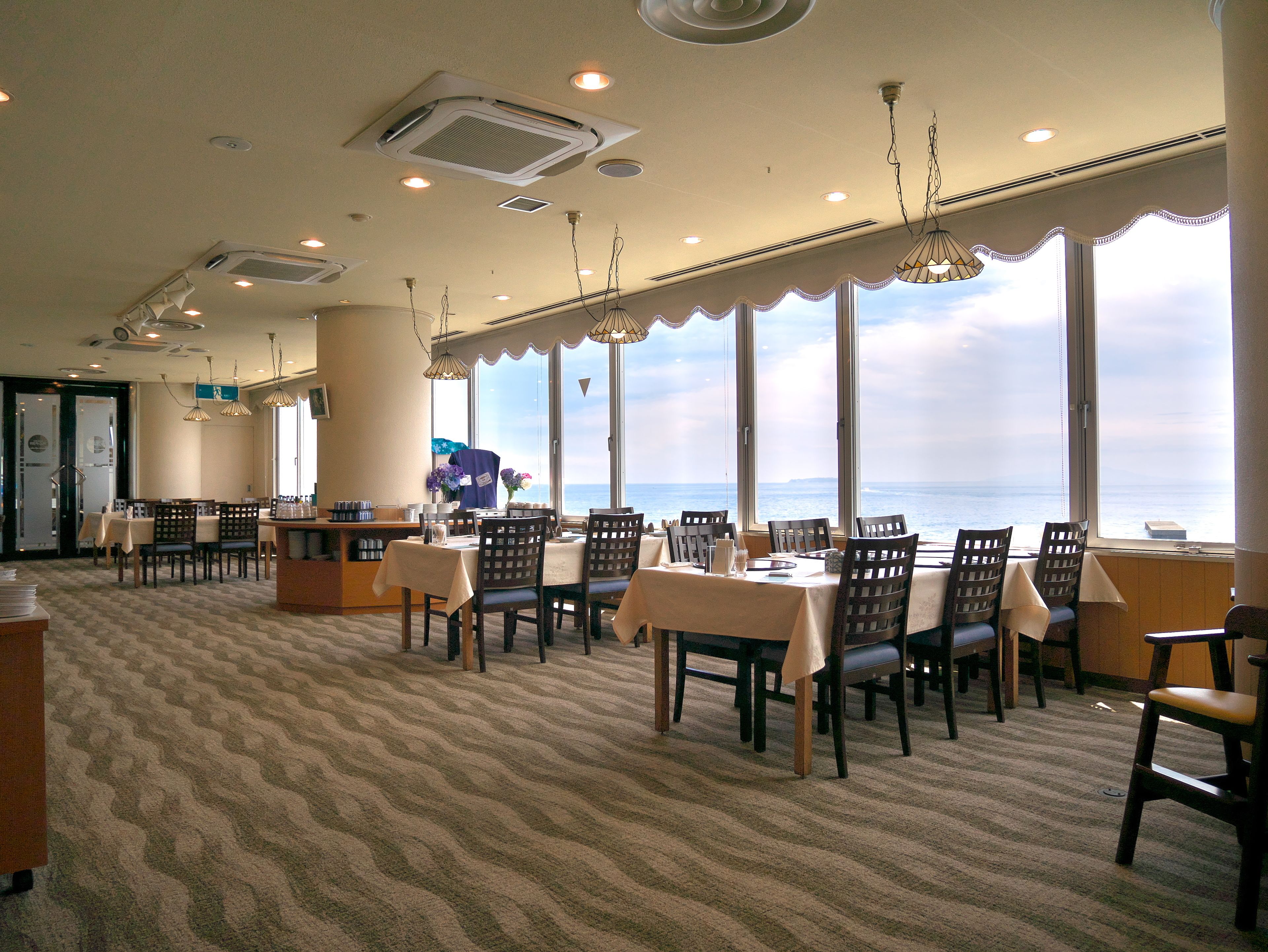 A dining area with tables and chairs, large windows with a view of the ocean, and ceiling vents.