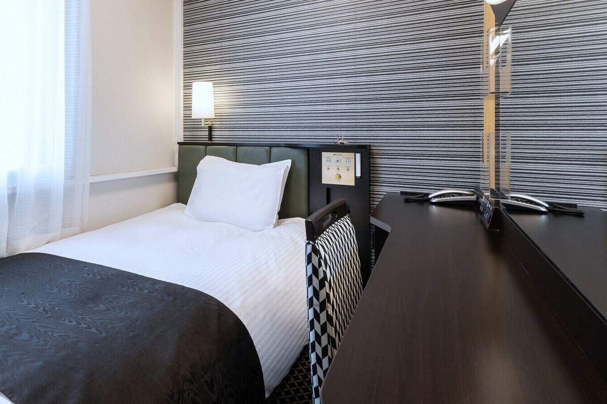 Habitación de hotel con cama, escritorio, silla, lámpara y panel de control en la pared.