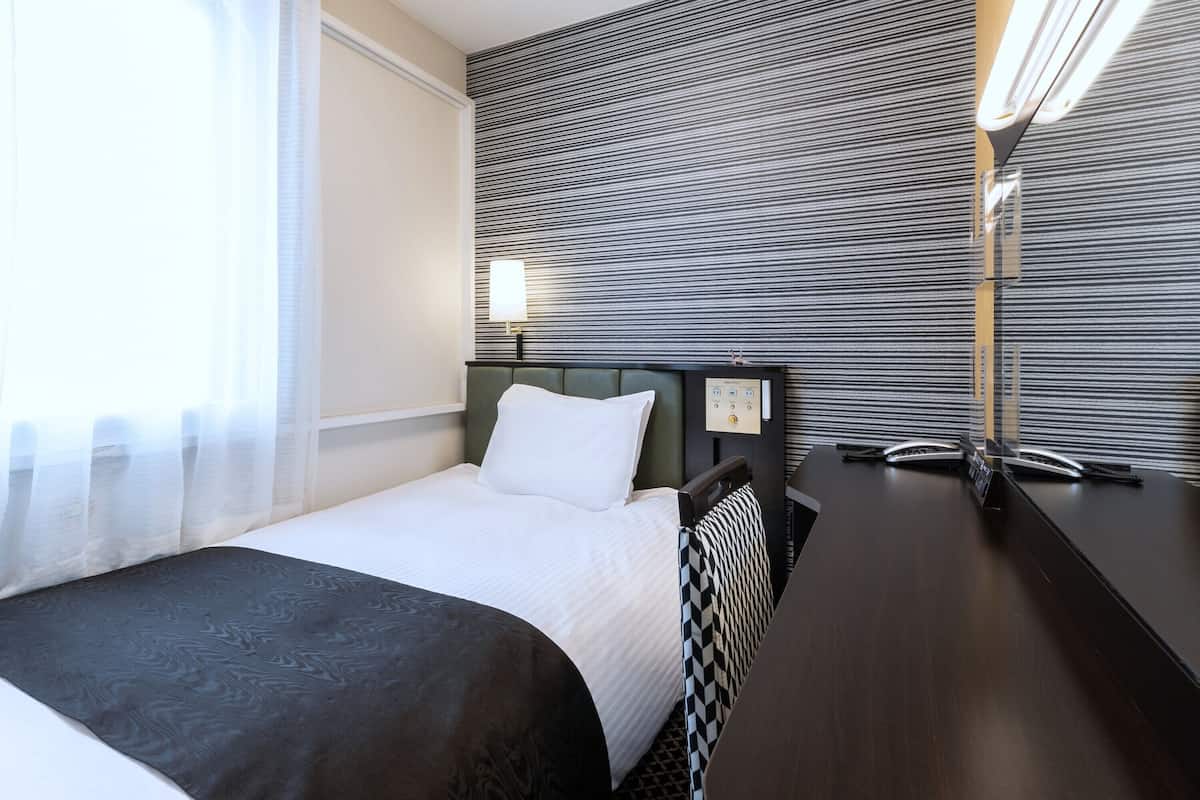 Habitación de hotel con cama, escritorio, silla, lámpara y panel de control en la pared.