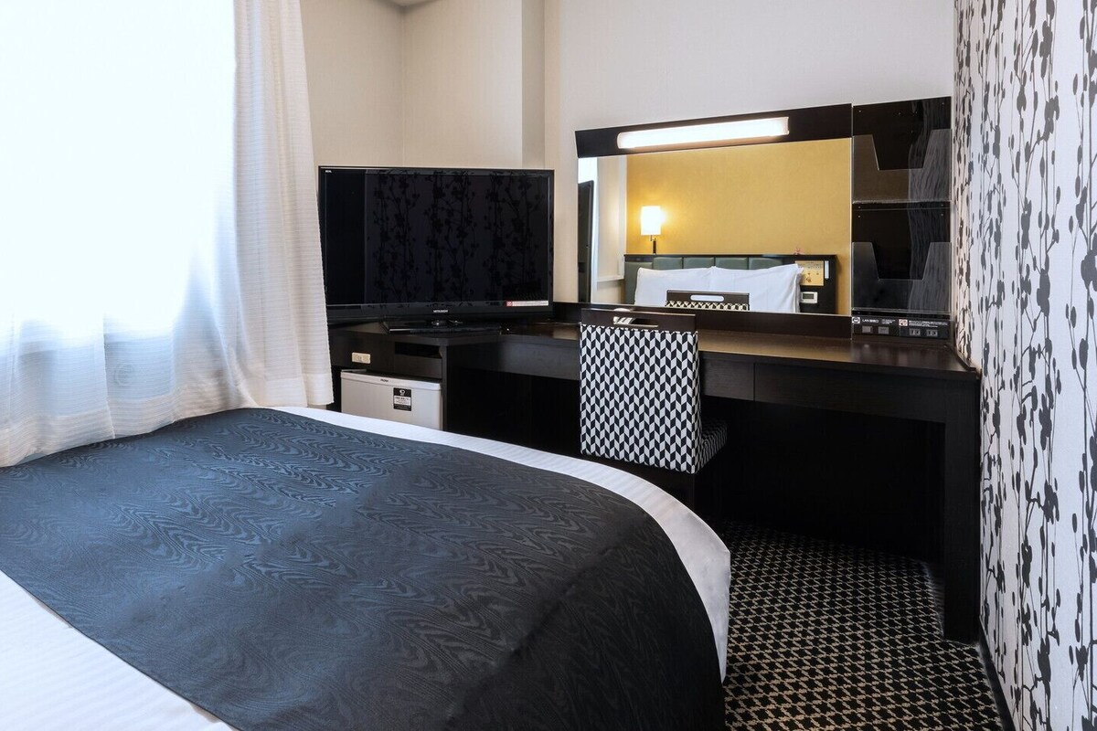 Habitación de hotel con cama, escritorio, silla, espejo y una pequeña kitchenette.