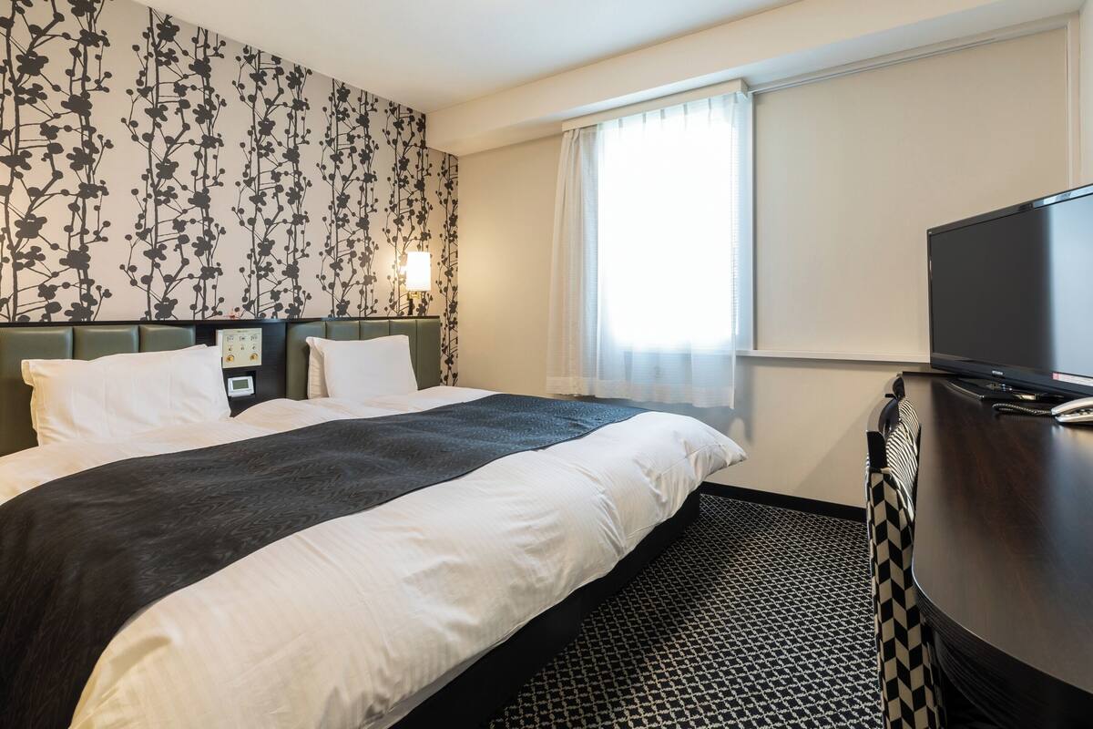 Una habitación de hotel con cama, escritorio, televisor y ventana con cortinas.