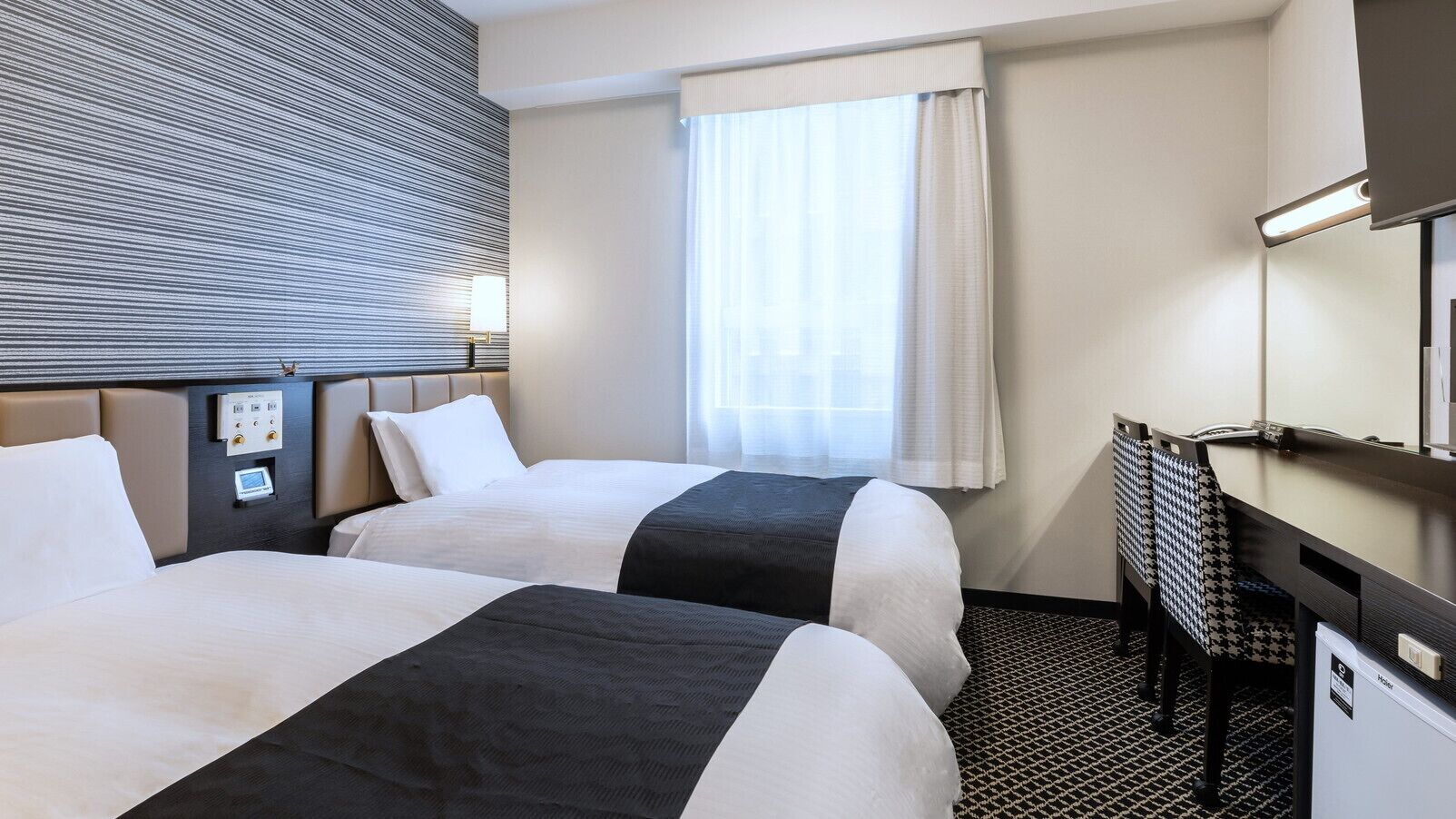 A hotel room with two beds, a desk, a chair, a window with curtains, and a wall-mounted control panel.