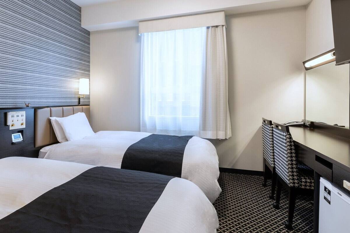 Habitación de hotel con dos camas, un escritorio, una silla, un ventanal con cortinas y un panel de control en la pared.
