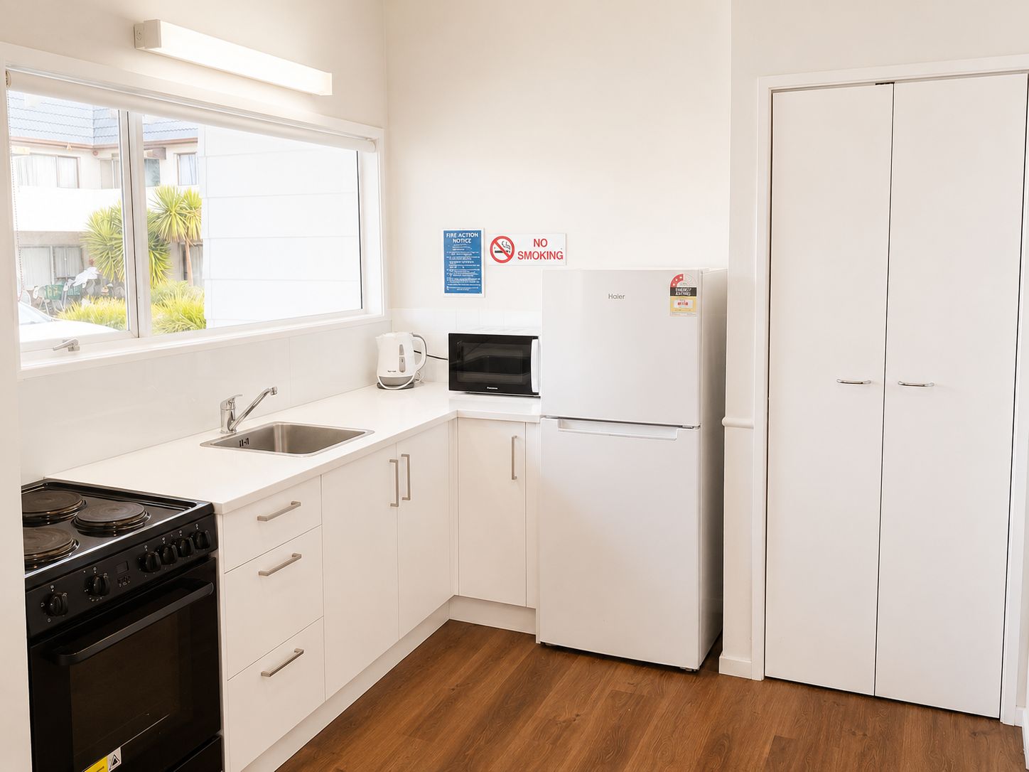 A kitchen with white cabinets, a stove, a microwave, and a refrigerator. There is a window with a view of the outside, and a door with a 'No Smoking' sign.