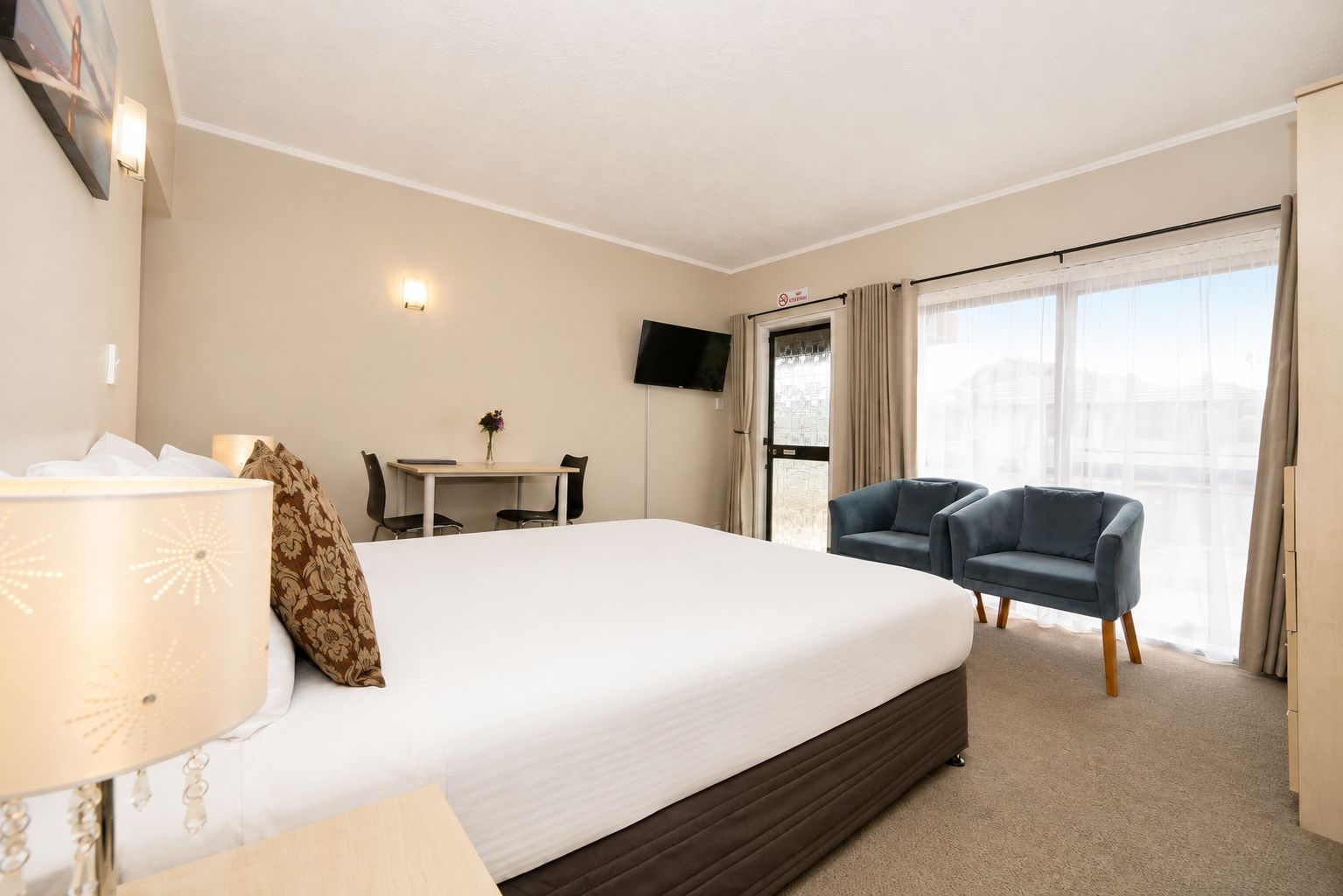 Habitación de hotel con cama, escritorio, dos sillas, televisor y ventana grande con cortinas.