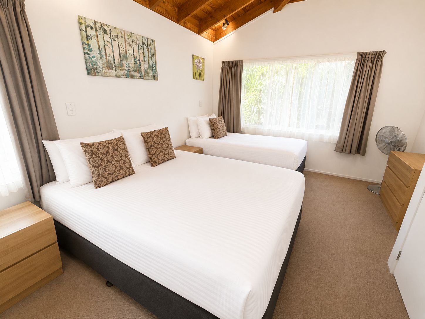 Une chambre d’hôtel avec un grand lit, deux tables de chevet, une commode en bois, un ventilateur de plafond et une fenêtre avec des rideaux.
