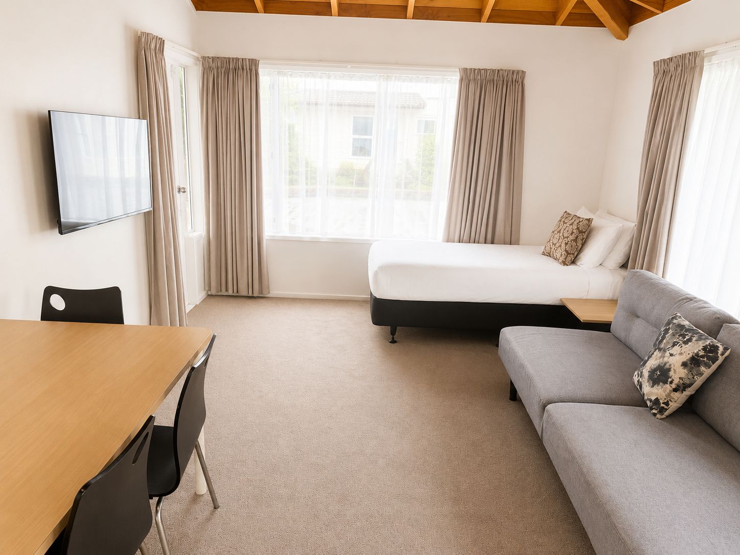 A hotel room with a bed, a sofa, a TV mounted on the wall, and a dining table with chairs.