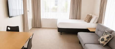 A hotel room with a bed, a sofa, a TV mounted on the wall, and a dining table with chairs.