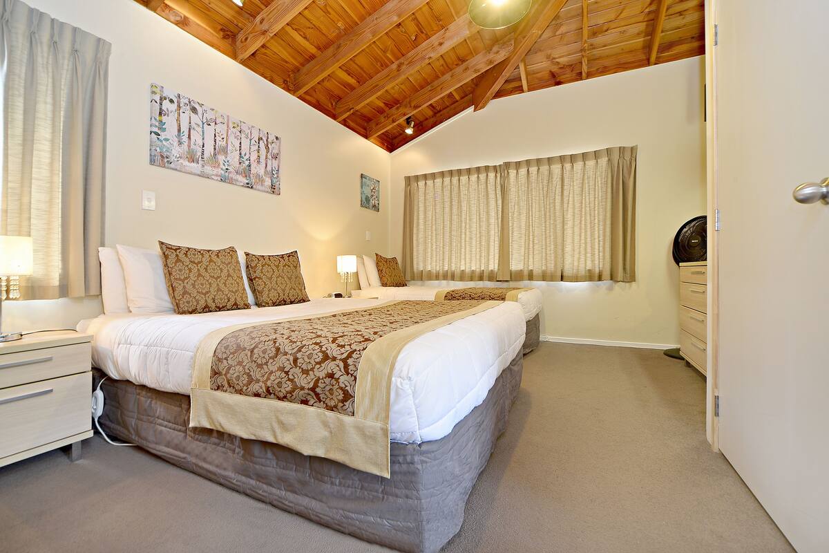 A bedroom with two beds, wooden ceiling beams, a large window with curtains, and a bedside table with a lamp.