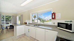 A kitchen with white cabinets, a dishwasher, a microwave, and a window with a view of palm trees and houses. - Takapuna Motor Lodge (Auckland)