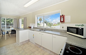 A kitchen with white cabinets, a dishwasher, a microwave, and a window with a view of palm trees and houses. - Takapuna Motor Lodge (Auckland)