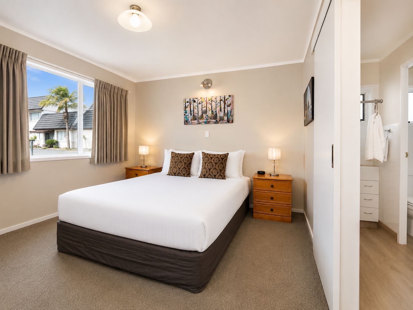 A hotel room with a large bed, two bedside tables with lamps, a window with curtains, a painting on the wall, and a bathroom visible through an open door.