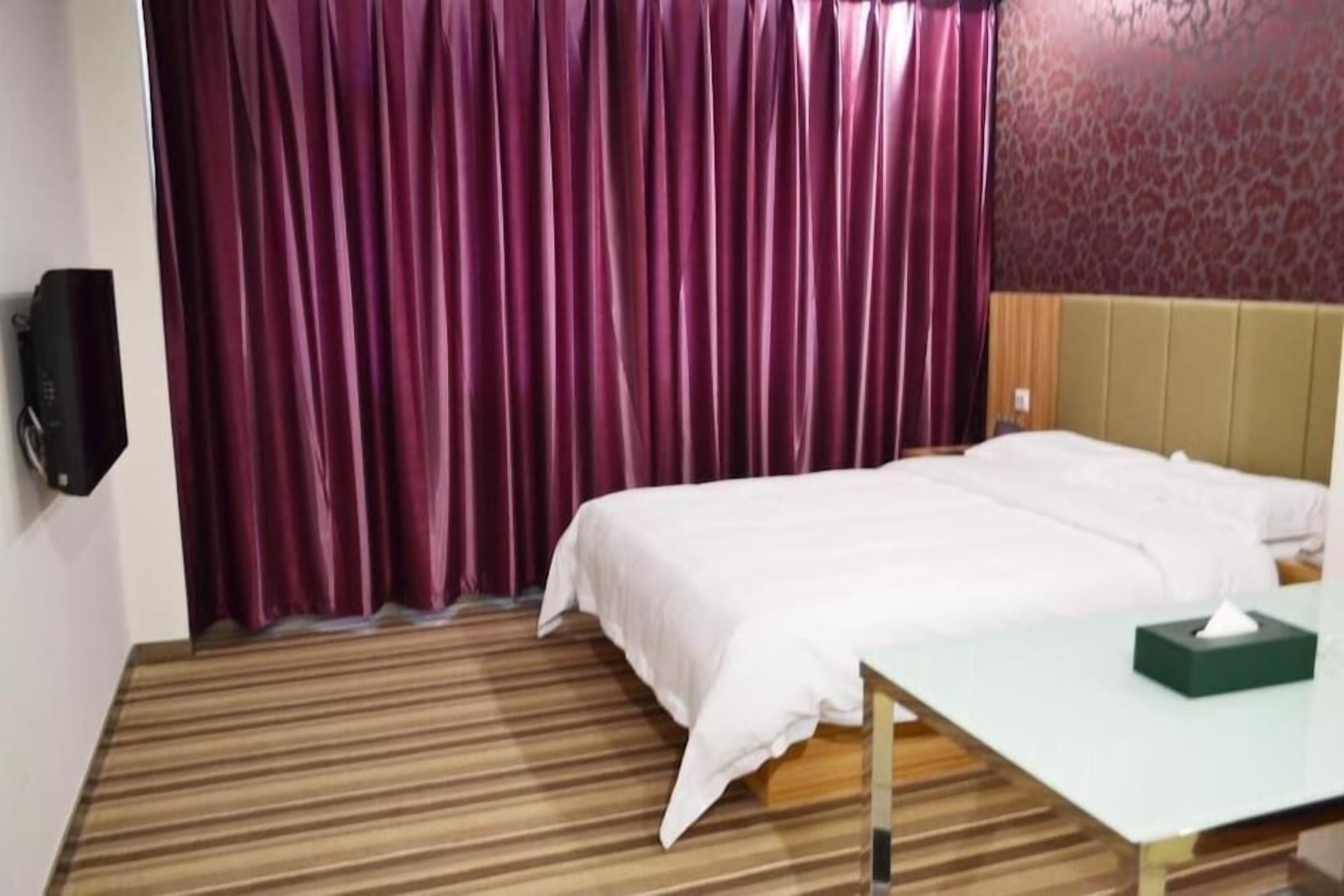 A hotel room with a bed, a bedside table, a green tissue box, and a wall-mounted speaker.