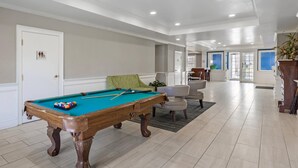 Una mesa de billar en una sala bien iluminada con una puerta señalizada para accesibilidad, una silla acolchada verde y vistas a otra sala con zona de comedor.