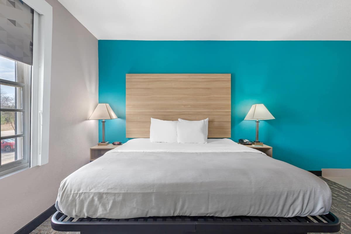 A hotel room with a bed, two bedside lamps, a wooden headboard, and a window with a view of a parking lot.