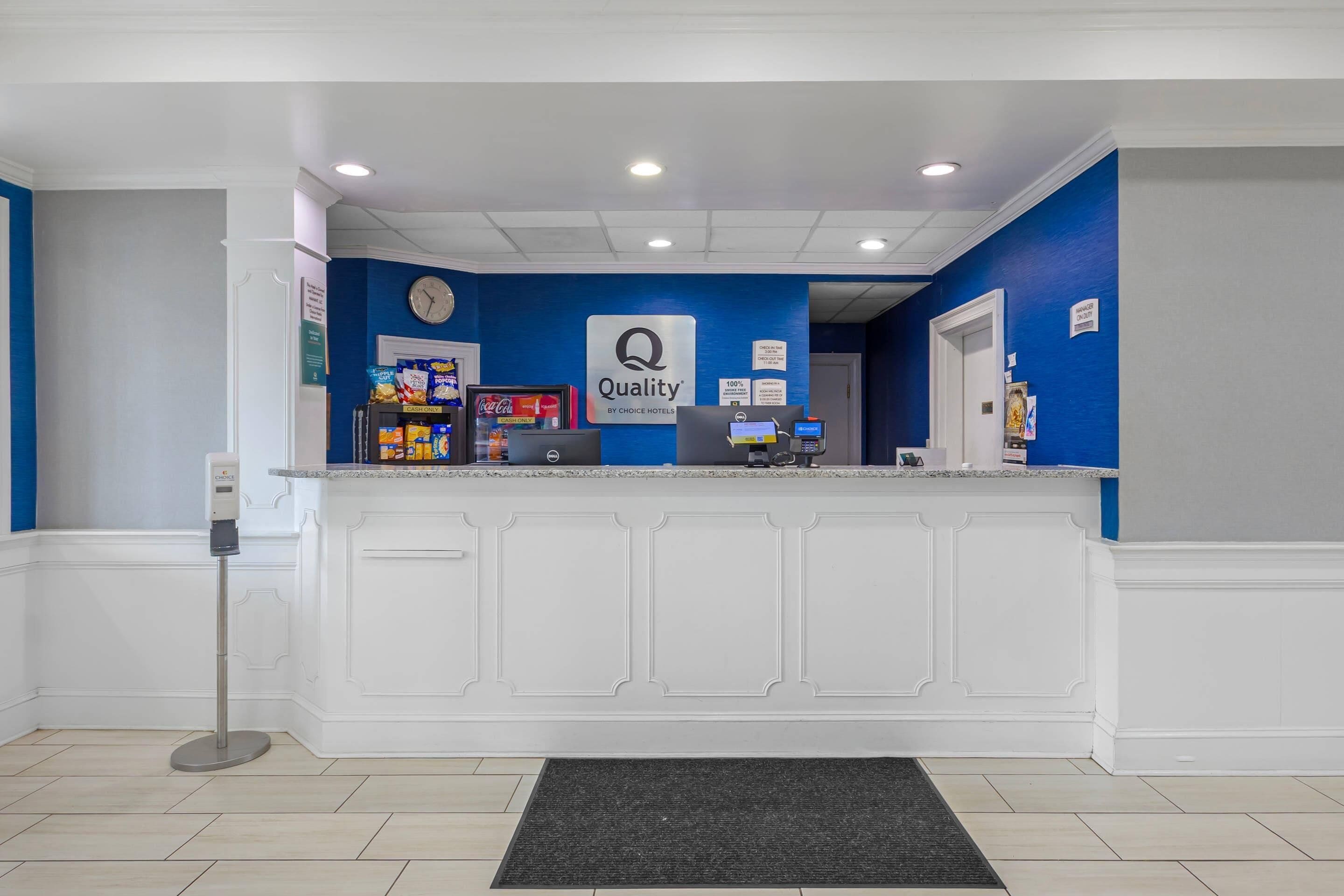 Un'area reception di un hotel con una parete blu, una sveglia, un cartello che recita 'Quality by Choice Hotels' e un bancone con diversi oggetti.