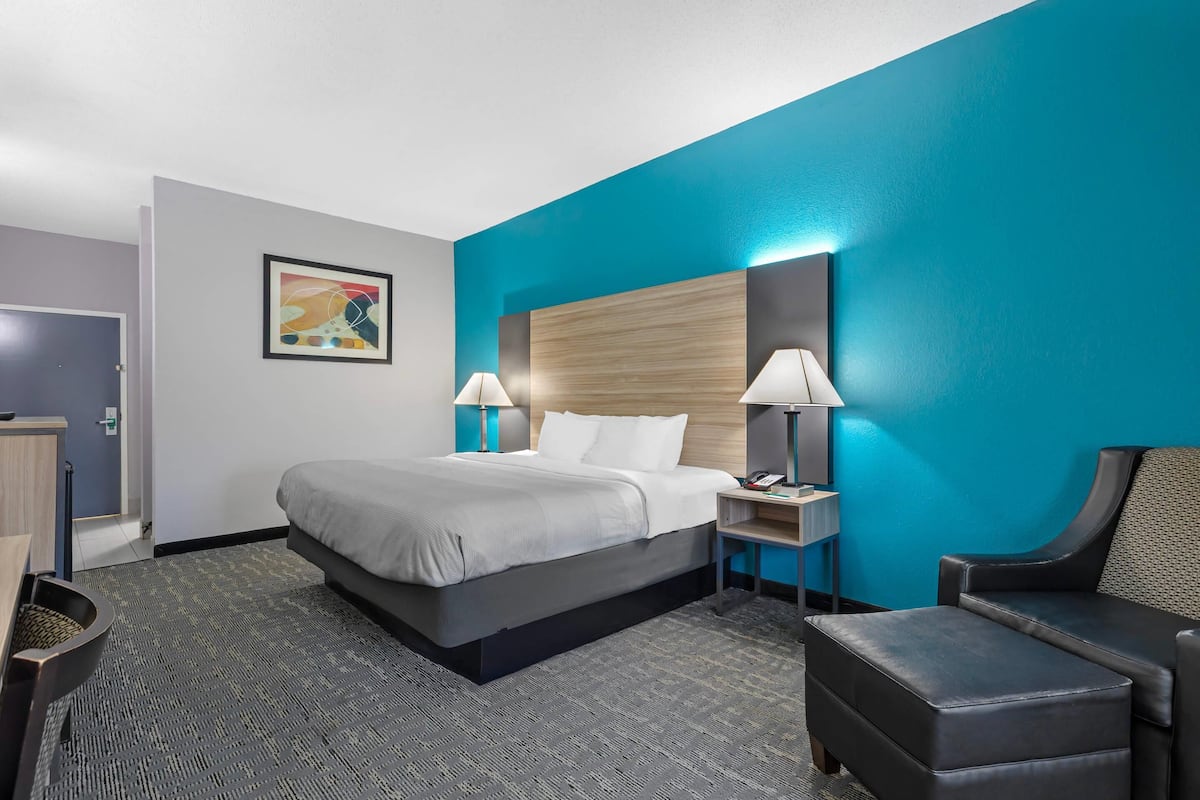 A hotel room with a bed, bedside lamps, a chair, a desk, and a painting on the wall.