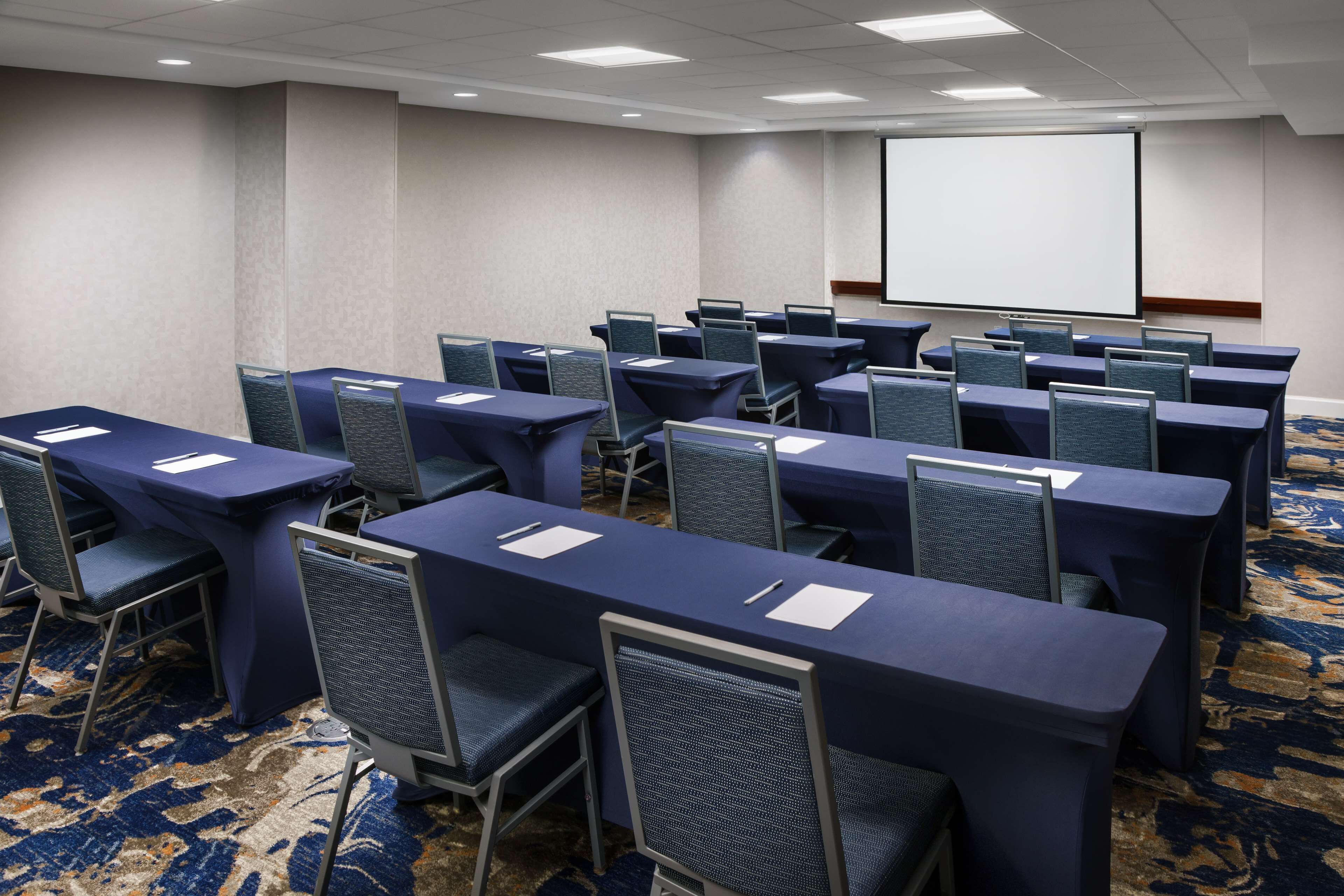 A conference room with rows of tables and chairs, a projector screen, and a carpeted floor.