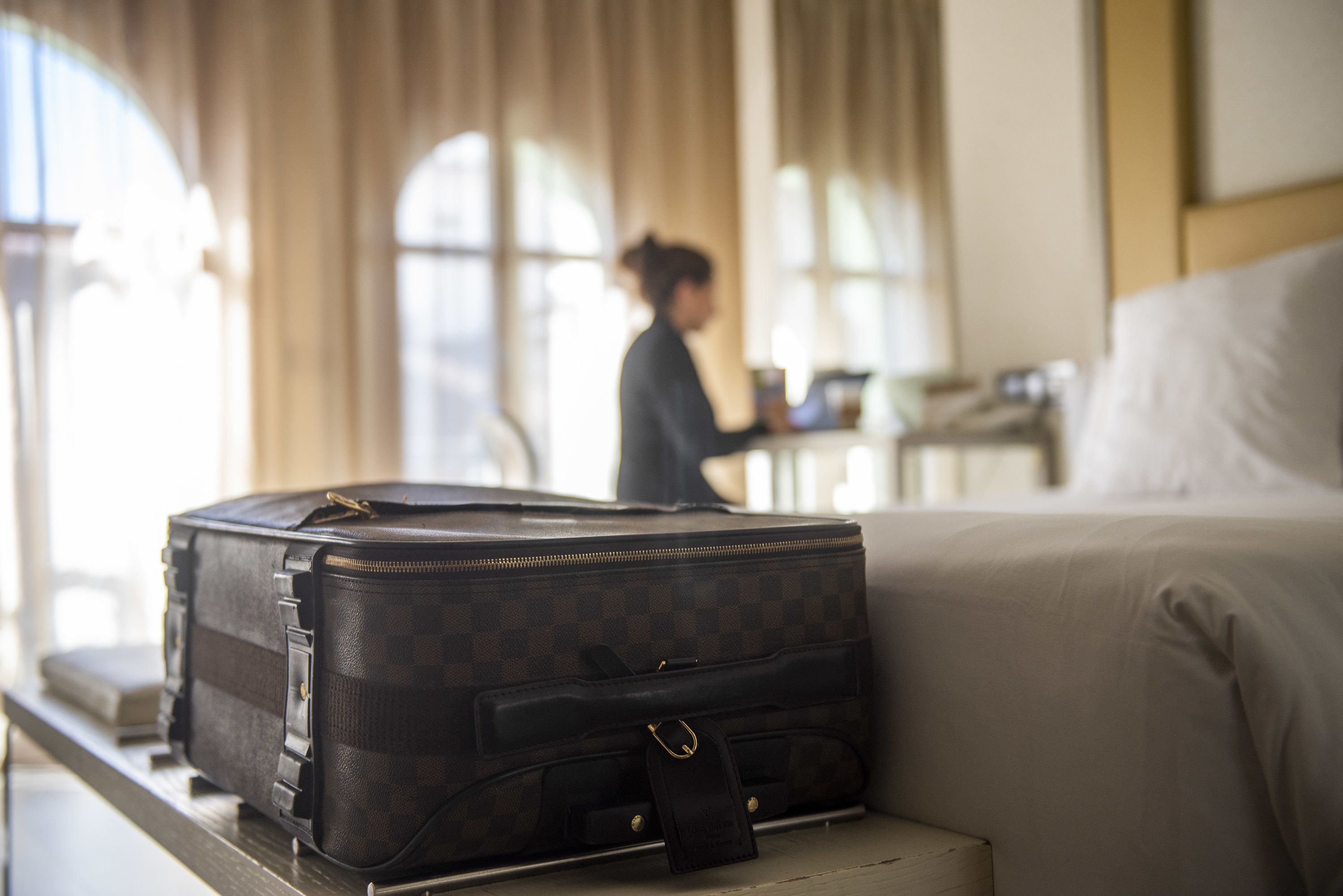 Une valise posée sur un lit dans une chambre d’hôtel, avec en arrière-plan une personne travaillant à un bureau.