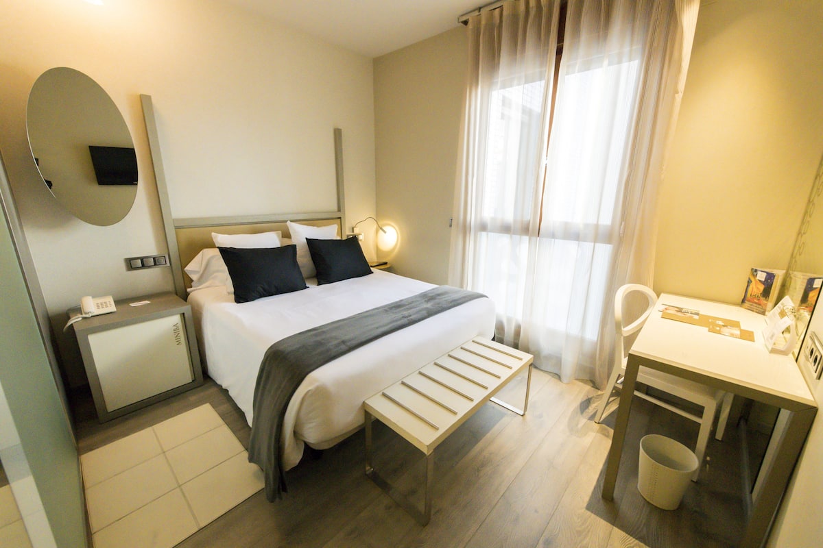 Ein Hotelzimmer mit Bett, Nachttisch, Schreibtisch, Stuhl und einem kleinen Mülleimer.