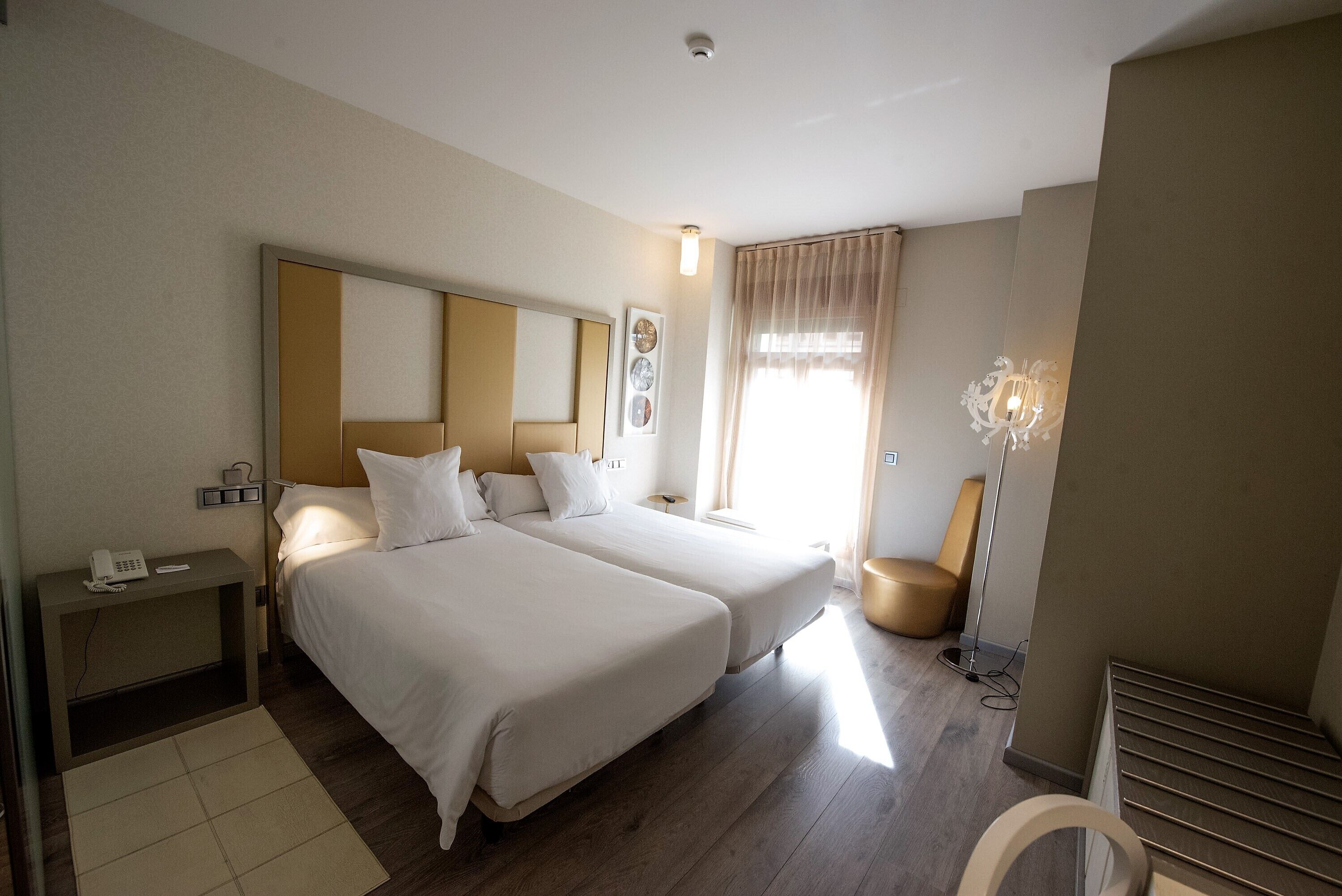 A hotel room with two beds, a bedside table with a phone, a lamp, and a window with curtains.