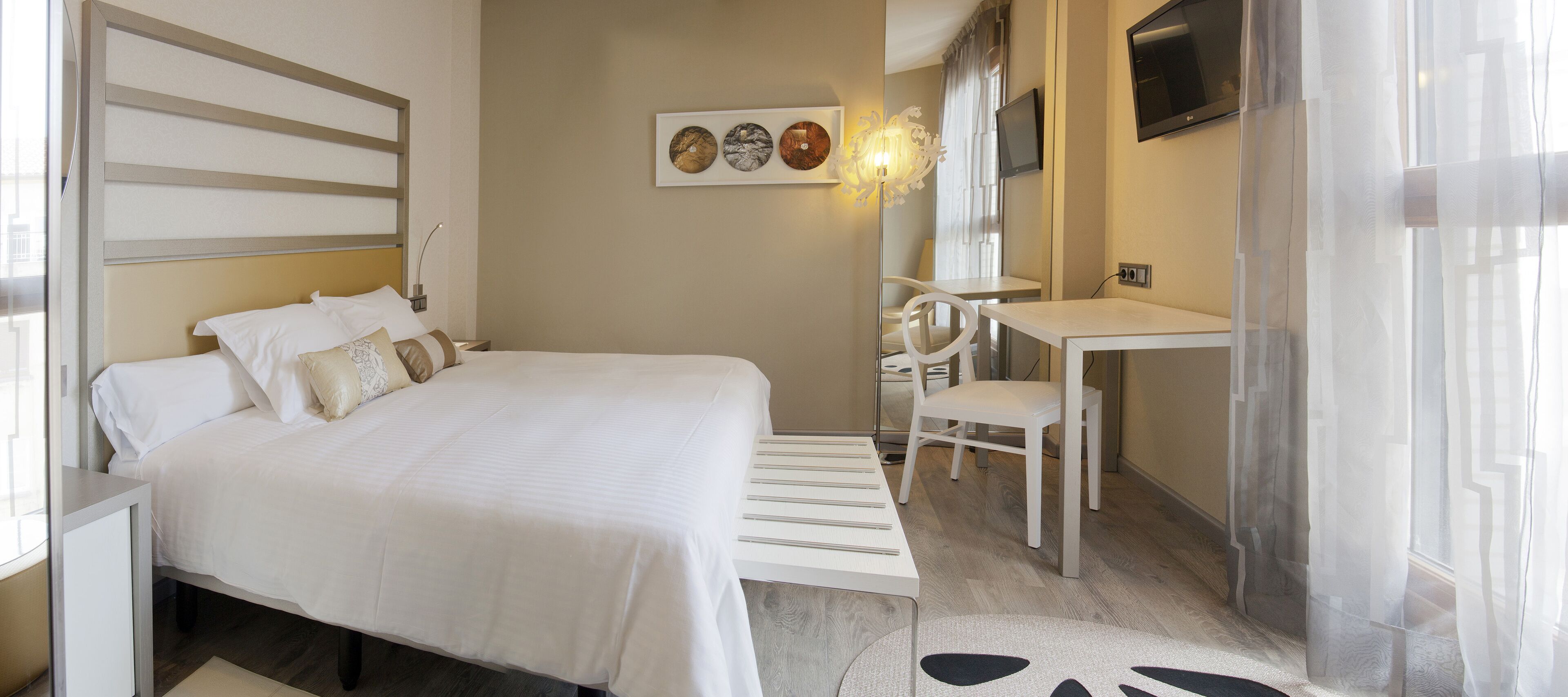 A hotel room with a bed, a desk, a chair, a TV, and artwork on the wall.