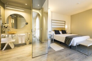 Una habitación de hotel moderna con un espejo grande, un lavamanos redondo, una mesita de noche, una cama con cabecero y un banco.