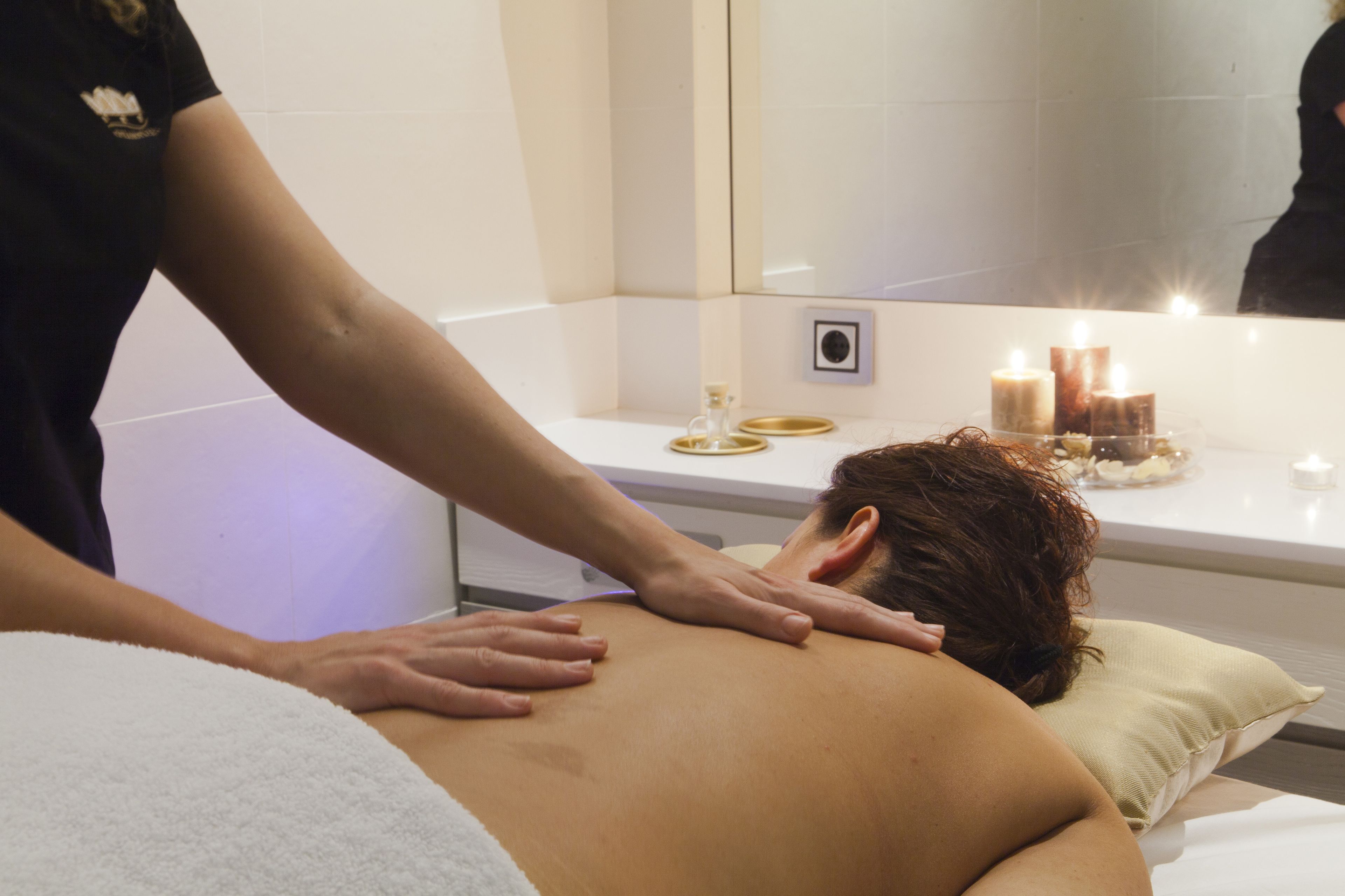 Une personne reçoit un massage dans une pièce où se trouvent un lit blanc, une petite table avec des bougies et un miroir qui reflète la scène.