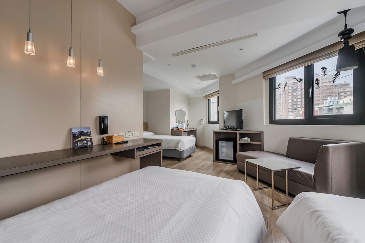 Une chambre d’hôtel moderne avec un grand lit, un canapé, un bureau et une télévision. On y trouve trois lampes suspendues et une fenêtre donnant sur des immeubles.