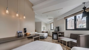 Una habitación de hotel moderna con una cama grande, un sofá, un escritorio y un televisor. Hay tres lámparas colgantes y una ventana con vistas a edificios.