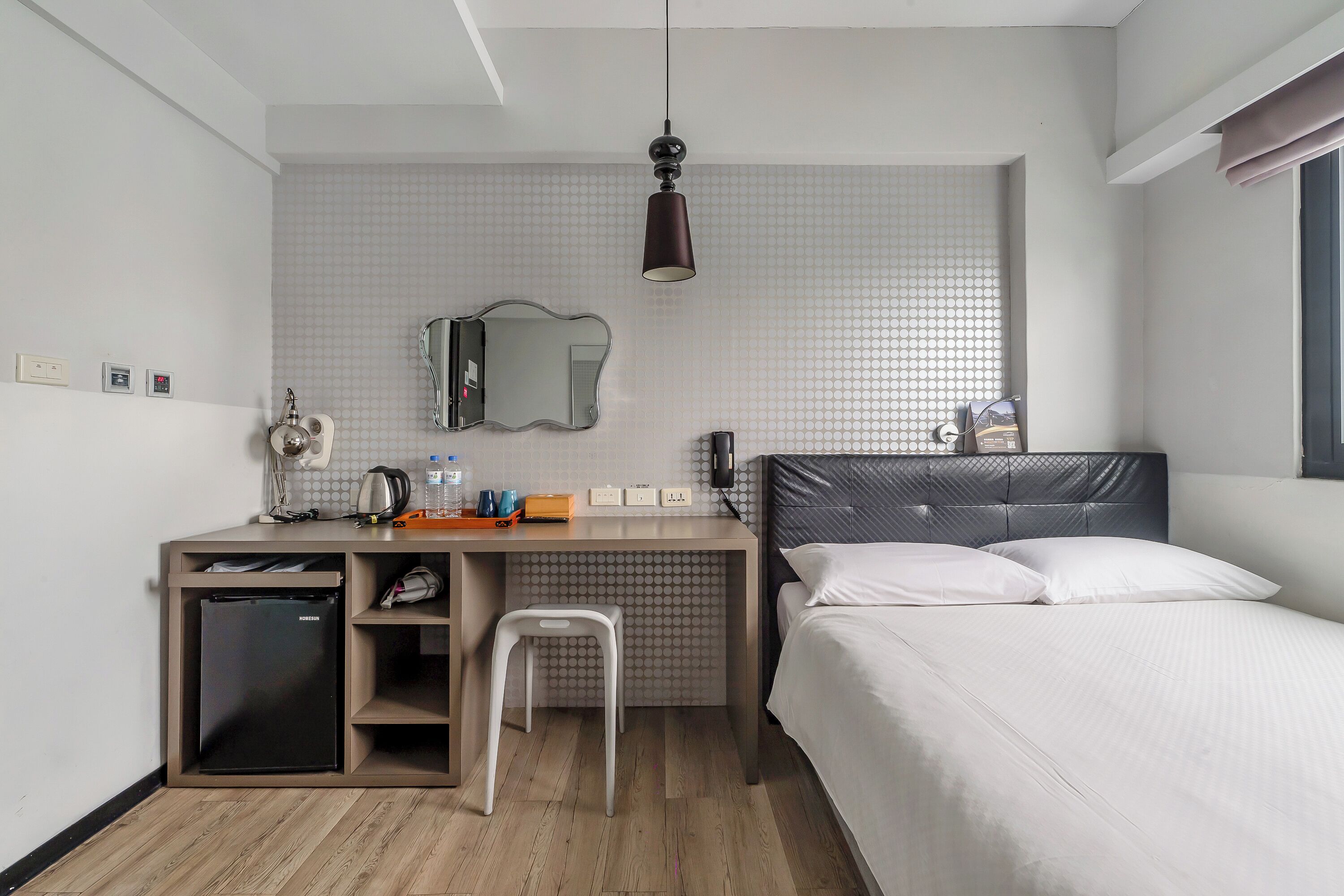 A compact hotel room with a bed, a desk with a chair, a small refrigerator, a mirror, and a hanging light fixture.